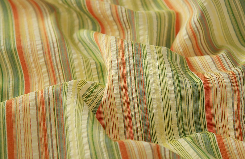 May include: Close-up of a striped fabric with orange, green, and white stripes. The fabric is woven and has a textured surface.