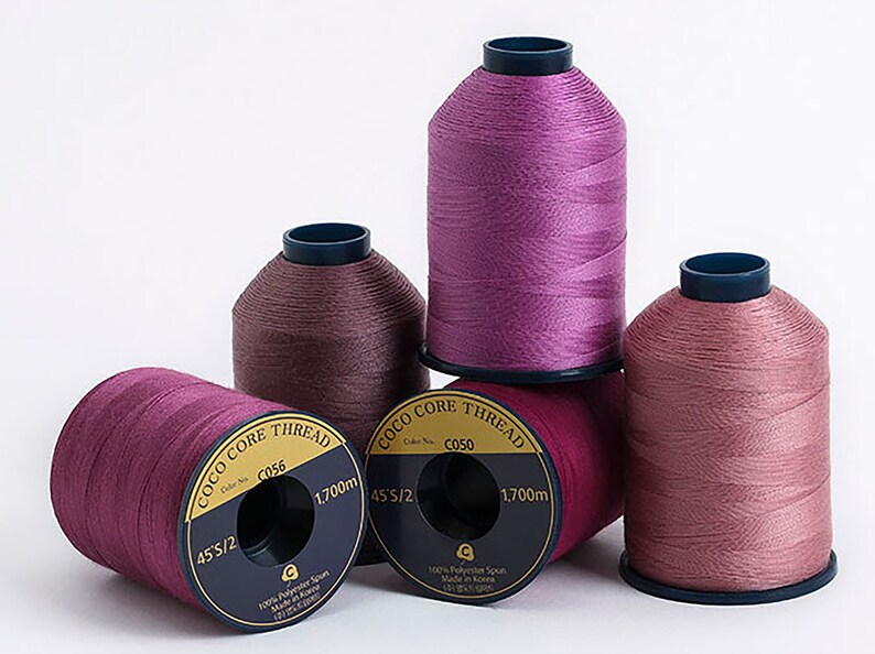 Puede incluir: Cuatro carretes de hilo rosa, malva y burdeos etiquetados "Coco Core Thread" con el texto "45'S/2", "CC050", "1.700 m" y "100% Polyester Spun Made in Korea".