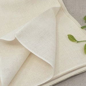 May include: A stack of off-white cotton fabric folded in a triangular shape. The fabric is smooth and has a subtle texture. A small green plant is in the background.