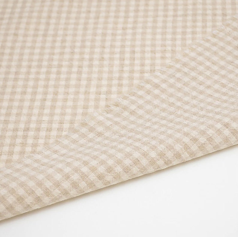 May include: A beige gingham fabric with a subtle texture. The fabric is folded in half, showing the pattern and weave.