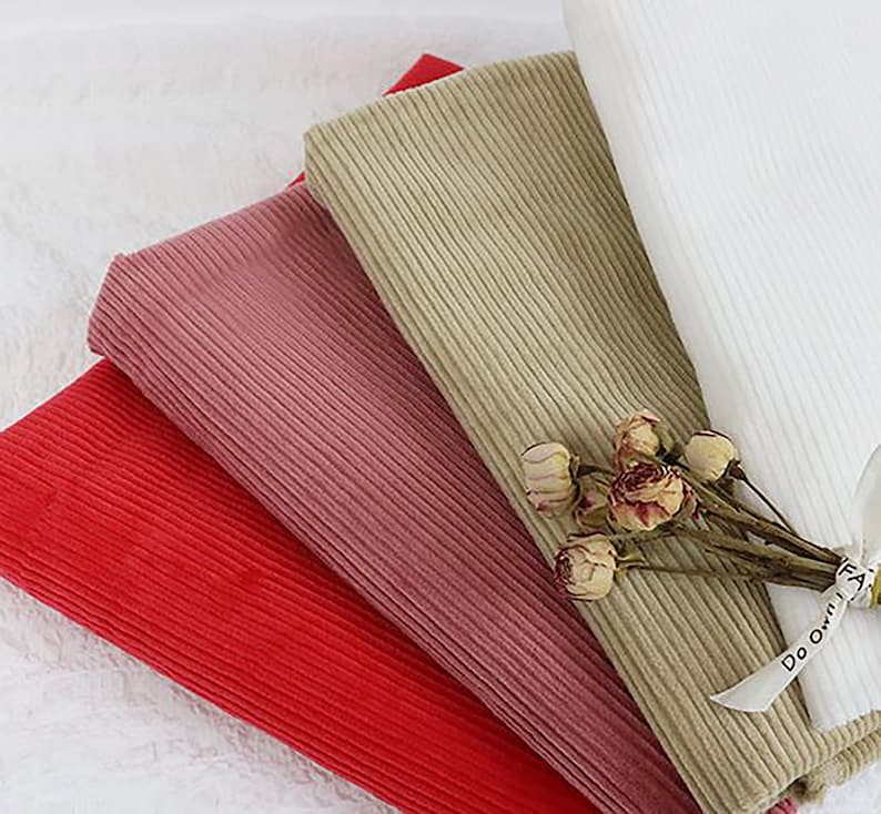 Op de afbeelding: Vier verschillende kleuren corduroy stofstalen. De kleuren zijn rood, roze, beige en wit. De stof is geribbeld en heeft een zachte textuur.