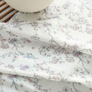 Könnte beinhalten: Weißer Stoff mit einem zarten Blumendruck mit kleinen rosa, lila und blauen Blüten auf dünnen Stielen. Der Stoff ist gefaltet und auf einer Holzoberfläche drapiert.