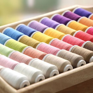 May include: A wooden tray containing a variety of thread spools in a spectrum of colours. The threads are in shades of blue, green, yellow, pink, orange, red, purple, and brown. The spools are arranged in neat rows, ideal for sewing or craft projects.