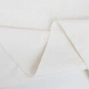 Op de afbeelding: Witte ribbelstof met een zacht, gestructureerd oppervlak. De stof is gevouwen in een driehoekige vorm, waardoor de textuur en de drape worden getoond.