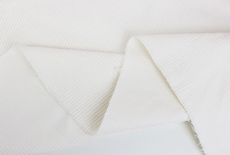 Op de afbeelding: Witte ribbelstof met een zacht, gestructureerd oppervlak. De stof is gevouwen in een driehoekige vorm, waardoor de textuur en de drape worden getoond.