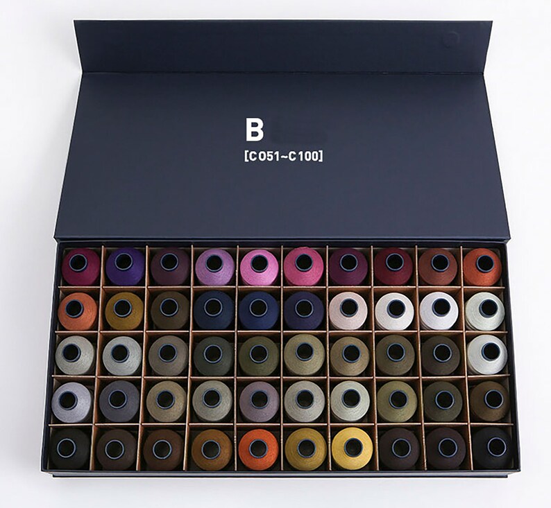 Puede incluir: Una caja negra con la tapa abierta, que revela una colecci&oacute;n de 50 carretes de hilo de varios colores. La caja est&aacute; etiquetada con "B [C 051 - C 100]".