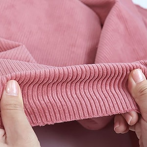 Op de afbeelding: Close-up van een roze corduroy stof met een geribbelde textuur. De stof wordt vastgehouden door een hand, waardoor de zachte en pluche textuur zichtbaar is.