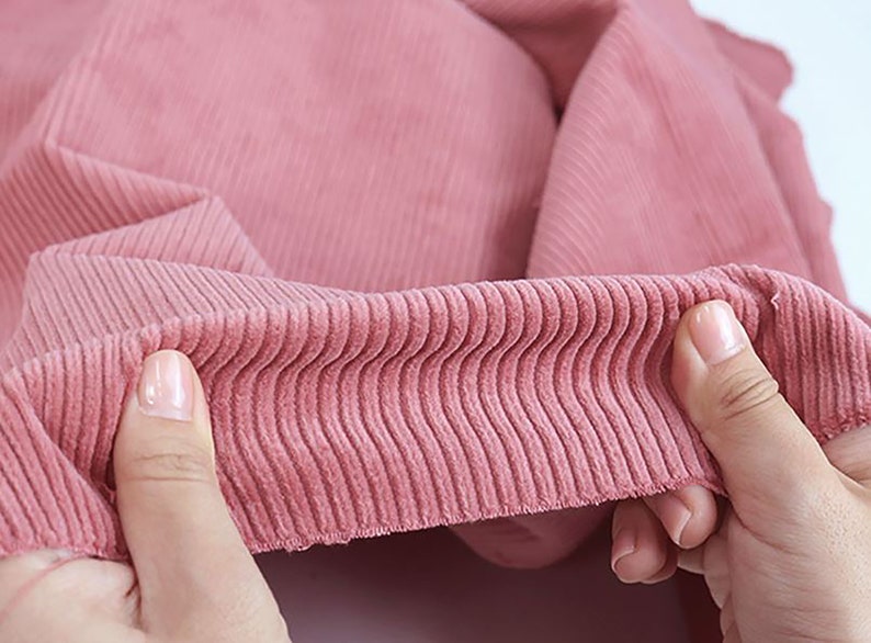 Op de afbeelding: Close-up van een roze corduroy stof met een geribbelde textuur. De stof wordt vastgehouden door een hand, waardoor de zachte en pluche textuur zichtbaar is.