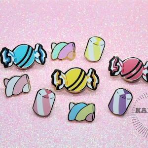Peut inclure: Plusieurs épingles en émail en forme de bonbons colorés. Les broches présentent des motifs de bonbons emballés en bleu, rose, jaune et violet, avec des contours dorés. Elles sont disposées sur un fond rose scintillant.