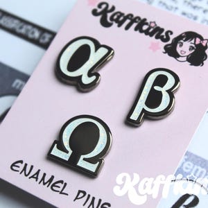 Pode incluir: Um conjunto de três pinos de esmalte com as letras gregas alfa, beta e ômega. Os pinos têm uma base preta com um contorno branco e brilhante. Os pinos são exibidos em um cartão rosa com o texto "ENAMEL PINS".