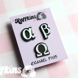 Pode incluir: Um conjunto de três pinos de esmalte preto e branco com as letras gregas alfa, beta e ômega. Os pinos são montados em um cartão lilás claro com o texto "ENAMEL PINS" e o nome da marca "Kaffkins".