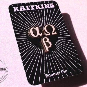 Pode incluir: Um alfinete de esmalte preto com as letras gregas alfa, ômega e beta em dourado. O alfinete está em um cartão preto com um design branco radiante e o texto "KAFFKINS". O cartão também tem o texto "Enamel Pin".