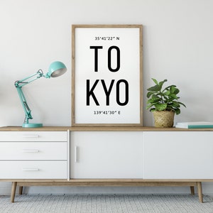 Puede incluir: Un cuadro enmarcado con la palabra "TOKYO" en letras negras grandes. La impresión incluye las coordenadas 35°41'22" N y 139°41'30" E. Se muestra en un mueble blanco y de madera con una lámpara de escritorio turquesa y plantas en macetas.