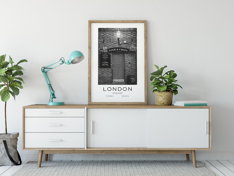 Puede incluir: Una impresi&oacute;n en blanco y negro enmarcada de una tienda de Fish & Chips de Londres, exhibida en un mueble blanco y de madera. Una l&aacute;mpara de escritorio turquesa y plantas en macetas flanquean la impresi&oacute;n. La impresi&oacute;n incluye el texto "LONDON ENGLAND".