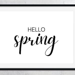 Puede incluir: Una impresión enmarcada con las palabras "HELLO SPRING" en escritura negra y fuente sans-serif sobre un fondo blanco. El marco es negro y la impresión se muestra contra una pared gris claro.
