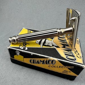 Peut inclure: Un rasoir de sécurité argenté vintage avec un manche strié repose sur une boîte noire et jaune. La boîte porte l'inscription "CHIAMCO COLLECTION" et un dessin d'homme. La tête du rasoir est visible.