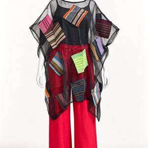 May include: A sheer black poncho featuring various colorful, rectangular fabric patches with striped patterns. The poncho is layered over a black tank top and bright red wide-leg trousers. The patches display diverse colors including pink, green, blue, and orange.