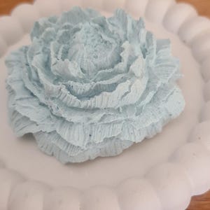 Könnte beinhalten: Ein hellblaues, blütenförmiges Deko-Objekt liegt auf einem weißen Teller mit gewelltem Rand. Die Blume hat detaillierte, geschichtete Blütenblätter. Der Teller steht auf einer Holzoberfläche. Dies ist wahrscheinlich ein Dekorationsartikel für das Zuhause.