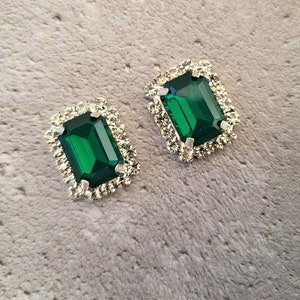 May include: A pair of emerald-cut earrings featuring vibrant emerald-green gemstones. Each earring is framed in silver and surrounded by small, clear crystals. The background is a light grey, textured surface.