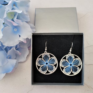 Op de afbeelding: Een paar zilverkleurige bloemenoorbellen met blauwe draadcentra, gepresenteerd in een zwarte doos. De oorbellen hebben een cirkelvormig ontwerp met een bloemenpatroon. Blauwe bloemen en een zilveren doos op de achtergrond.