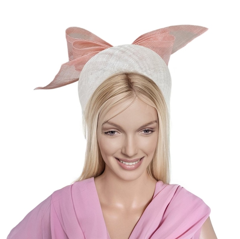 May include: A white and pink hat with a large bow. The hat is made of a straw-like material and has a wide brim. The bow is made of a soft, pink fabric.