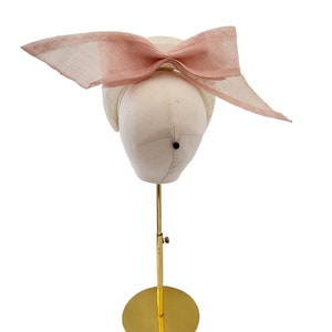 May include: A white hat with a large pink bow on top. The hat is displayed on a gold stand.