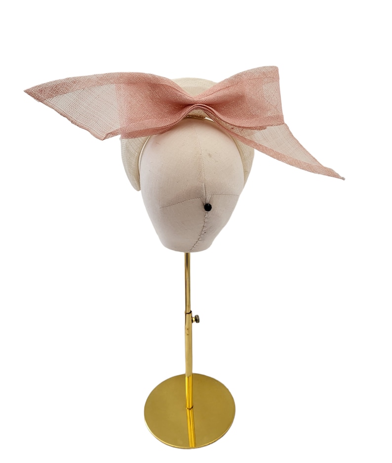 May include: A white hat with a large pink bow on top. The hat is displayed on a gold stand.