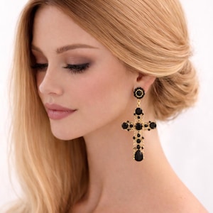 May include: A woman with blonde hair wearing gold-coloured cross earrings adorned with black gemstones. The earrings are a striking feature, hanging from the earlobes. The woman has fair skin and subtle makeup.