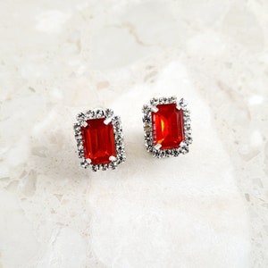 May include: A pair of red gemstone earrings with a silver setting and a clear crystal halo.