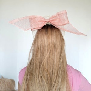 May include: A white hat with a large pink bow on top. The hat is worn on a person with long blonde hair.