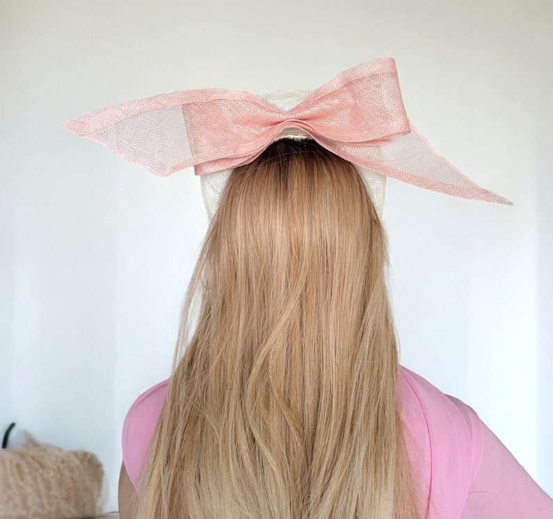 May include: A white hat with a large pink bow on top. The hat is worn on a person with long blonde hair.