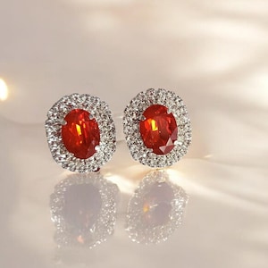 Può includere: Un paio di orecchini ovali con una pietra rossa e un alone di pietre bianche più piccole.