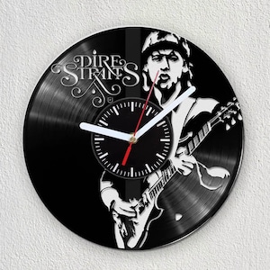 May include: A black vinyl record clock featuring a silhouette of a musician playing a guitar. The clock face is white with black numbers and hands. The record has the text "Dire Straits" printed on it.