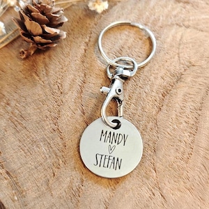 Könnte beinhalten: Ein kreisförmiger Schlüsselanhänger aus silberfarbenem Metall mit Karabinerverschluss und Schlüsselring. Der runde Anhänger ist mit dem Text MANDY, einem kleinen Herzsymbol und dem Namen STEFAN in einer schlichten, schwarzen Schrift graviert.