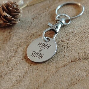 Op de afbeelding: Zilveren sleutelhanger met een rond label gegraveerd met de namen "Mandy" en "Stefan" gescheiden door een hart symbool.