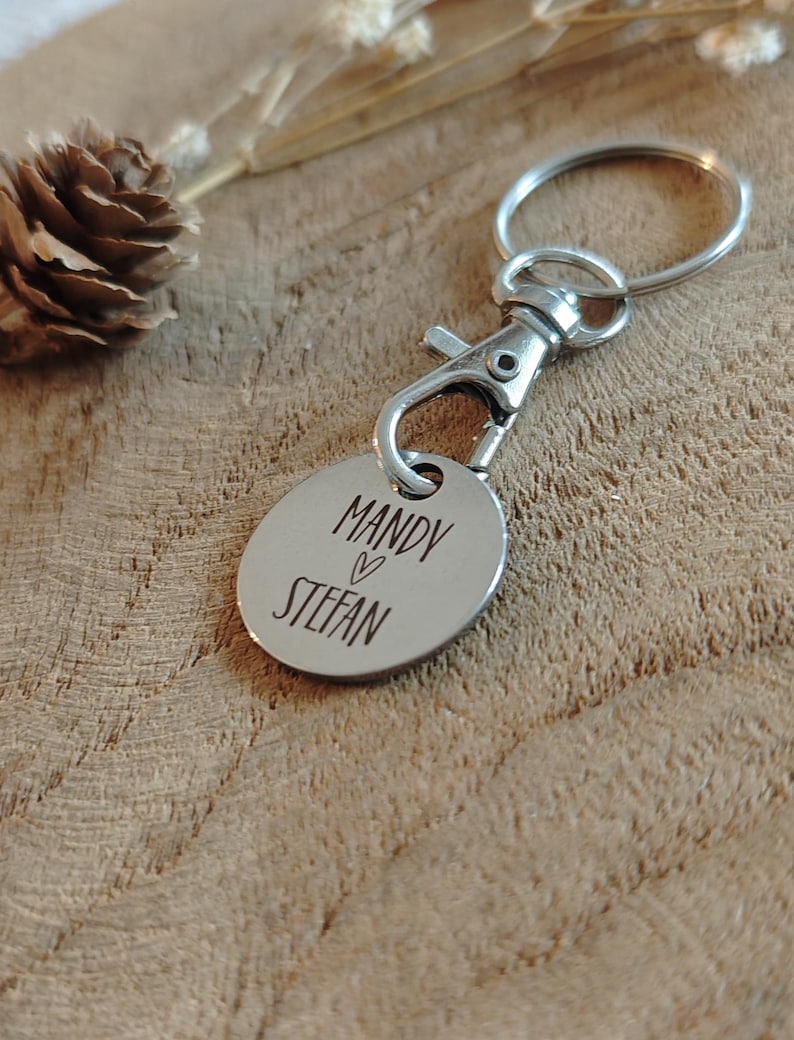 Op de afbeelding: Zilveren sleutelhanger met een rond label gegraveerd met de namen "Mandy" en "Stefan" gescheiden door een hart symbool.