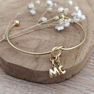 Könnte beinhalten: Goldfarbenes Armreif-Armband mit Knoten-Design und hängenden Initialen "MC". Das Armband hat eine glatte, polierte Oberfläche und eine kleine Kugel am Ende. Das Armband wird auf einer Holzoberfläche präsentiert.