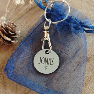 May include: A silver keychain with a round tag that says "JONAS" with a small heart symbol. The keychain is attached to a blue mesh bag.