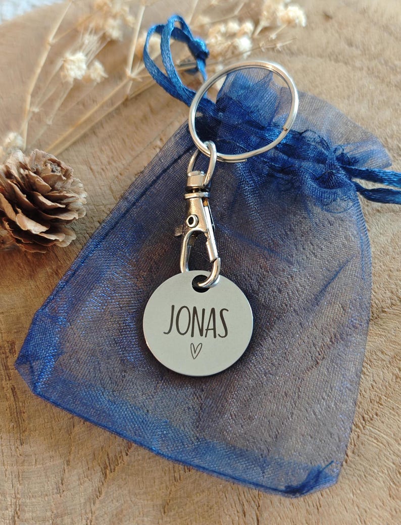 May include: A silver keychain with a round tag that says "JONAS" with a small heart symbol. The keychain is attached to a blue mesh bag.