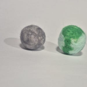 May include: Two felt balls, one grey and one green and white. The grey ball has a mottled appearance. The green and white ball has a design that resembles a globe. Both balls are round and sit on a white surface.
