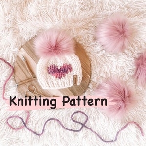 Puede incluir: Patrón de tejido para un gorro blanco con un diseño de corazón rosa y un pompón rosa esponjoso. El gorro está sobre una superficie de madera con hilo y agujas de tejer.