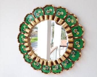 May include: A round mirror with a gold frame and green and white floral details. The mirror has a scalloped edge and is surrounded by a decorative border.