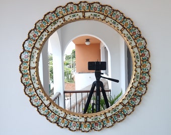 May include: A round, ornate mirror with a white background and gold trim. The mirror has a floral design with green, blue, and pink accents. The mirror is reflecting a room with a tripod and a camera.