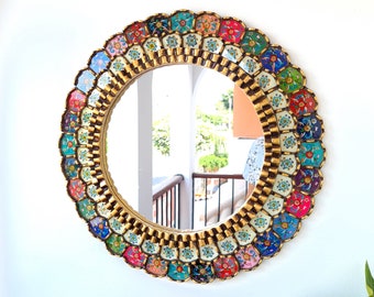 May include: Round decorative mirror with a gold frame and a colorful, mosaic-style border. The border features a repeating pattern of small, multi-colored tiles in shades of blue, red, green, and yellow. The mirror is hanging on a white wall.
