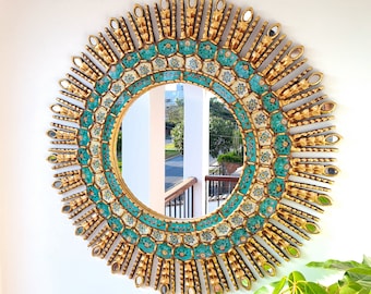 May include: A round decorative mirror with a gold frame and a turquoise and white mosaic border. The frame has a sunburst design with small, oval mirrors. The mirror is mounted on a white wall, with a glimpse of a balcony and greenery in the reflection.