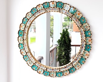 May include: Round decorative mirror with a gold frame and a scalloped edge. The frame is adorned with turquoise, white, and floral patterned tiles. The mirror reflects a view of a green tree and a white wall.