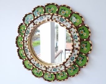 May include: Round decorative mirror with a scalloped, green and gold frame. The frame features floral designs in green, white, and blue. The mirror reflects a view of a balcony and trees.