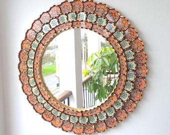 May include: A round mirror with a decorative frame made of painted wood. The frame is decorated with a floral pattern in shades of pink, blue, and green. The mirror is perfect for adding a touch of bohemian style to any room.