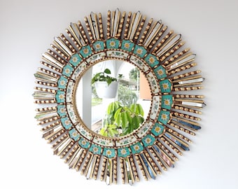 May include: A round sunburst mirror with a gold frame and turquoise and white mosaic tiles. The mirror reflects a potted plant and a window.