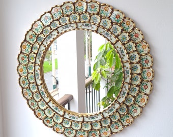 May include: A round mirror with a decorative frame made of white tiles with gold accents and floral designs. The tiles are arranged in a circular pattern around the mirror.
